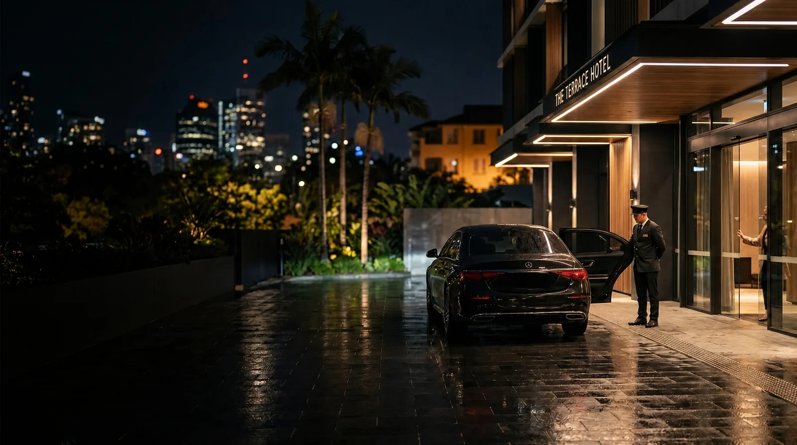 A chauffeur waiting beside a black sedan at a luxury hotel entrance on a rainy evening.